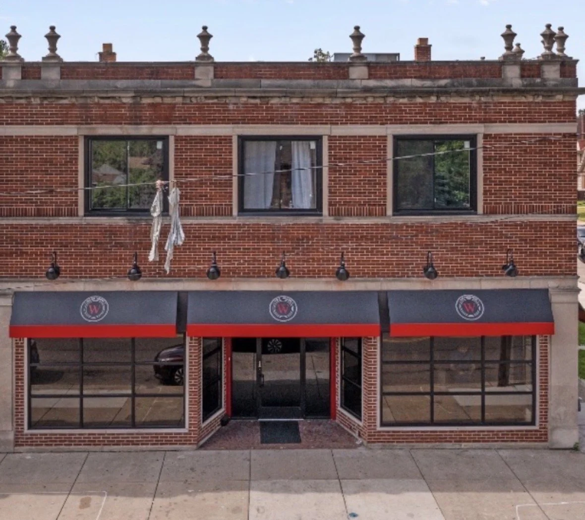 A brick building with three windows on the upper floor and two large glass storefront windows with an entrance in the center on the ground floor. A black and red awning with a logo extends over the storefront windows. A clothesline with laundry hangs