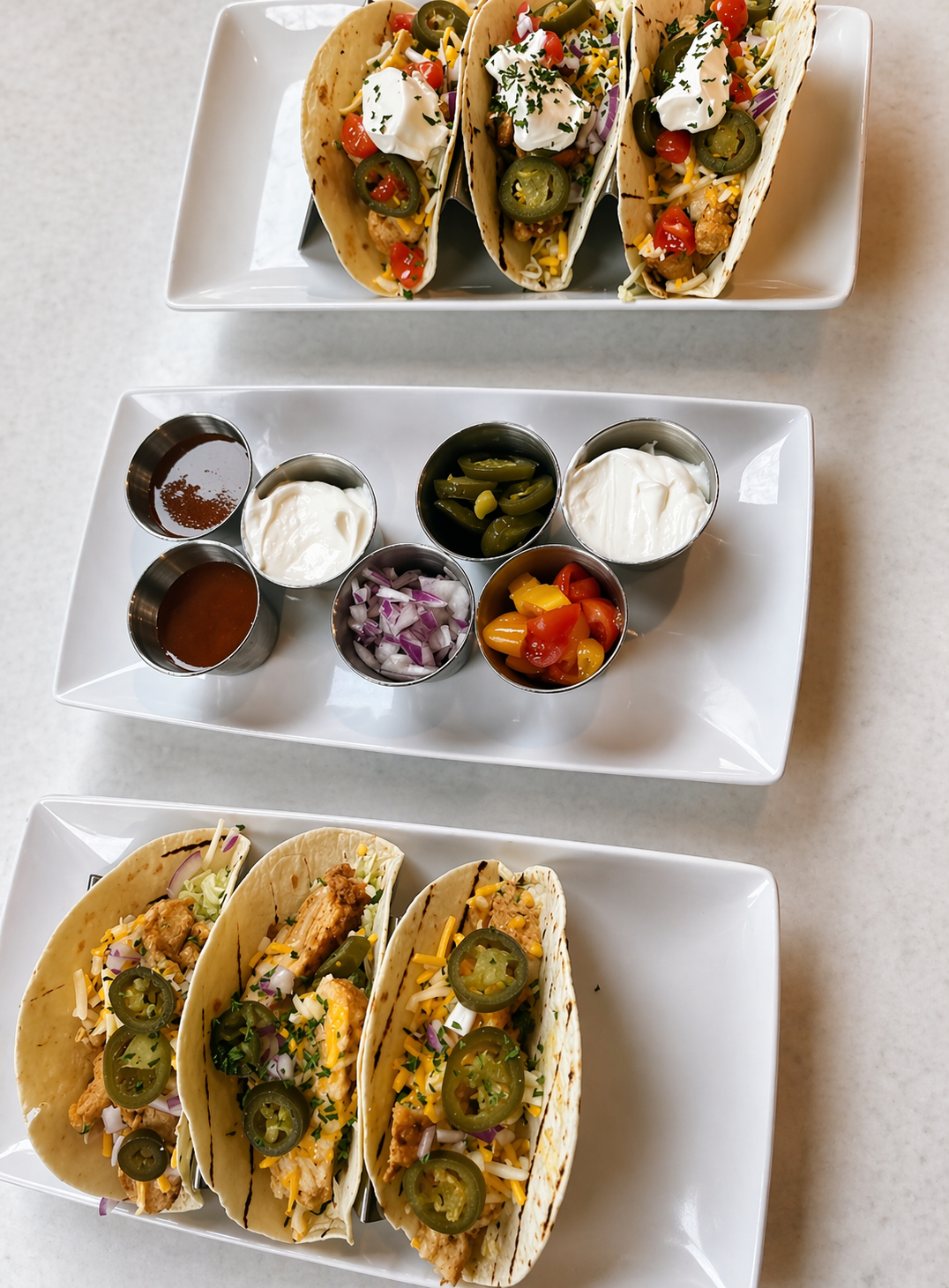 Three plates of tacos filled with chicken, shredded cheese, jalapeños, and toppings, served with various condiments and toppings including salsa, sour cream, shredded lettuce, chopped onions, and chopped tomatoes.