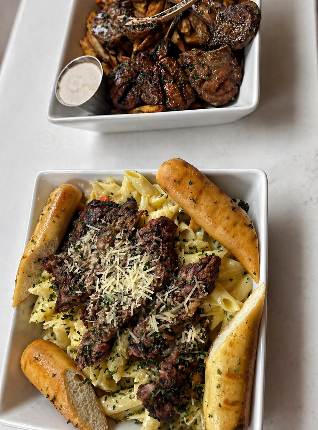 Two white square dishes of food. The top dish contains grilled lamb chops with herbs, grilled vegetables, and a side of dipping sauce. The bottom dish has pasta with grilled steak strips, grated cheese, and toasted bread slices.