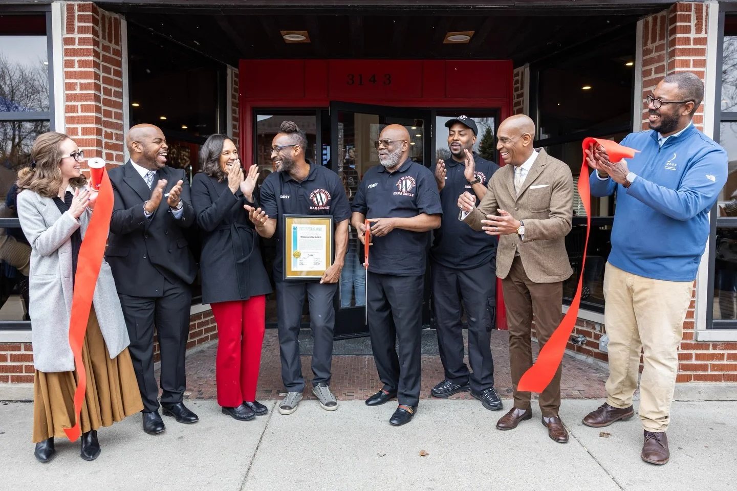 Group of people celebrating a ribbon-cutting ceremony outside a building. Some are clapping, a man is holding a framed certificate, and others are holding scissors dressed in business and casual attire.