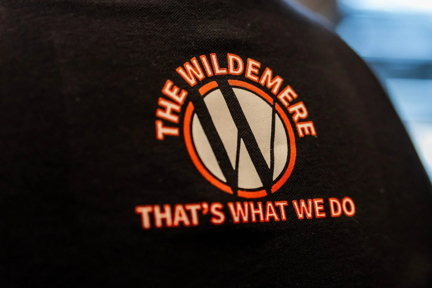 Close-up of a black fabric with an embroidered logo and text for 'The Wilderness That’s What We Do', featuring a stylized 'W' inside a circle.