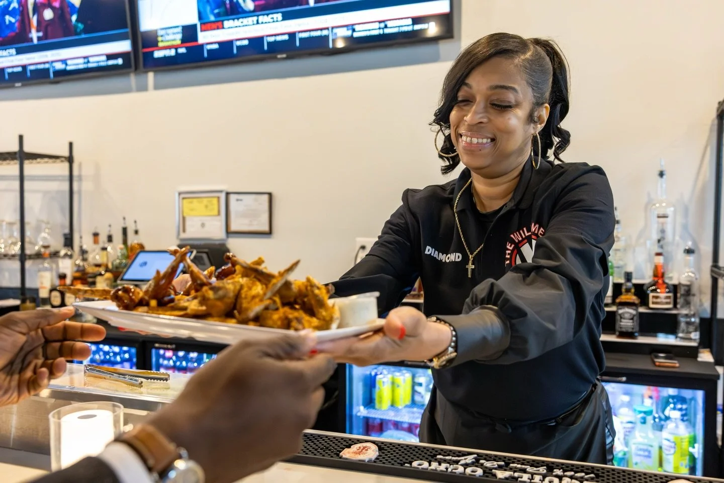 A woman at a bar is handing a plate of chicken wings to a customer. She is smiling and wearing a black shirt with a logo and name tag, in a setting with a TV screen and beverage bottles in the background.