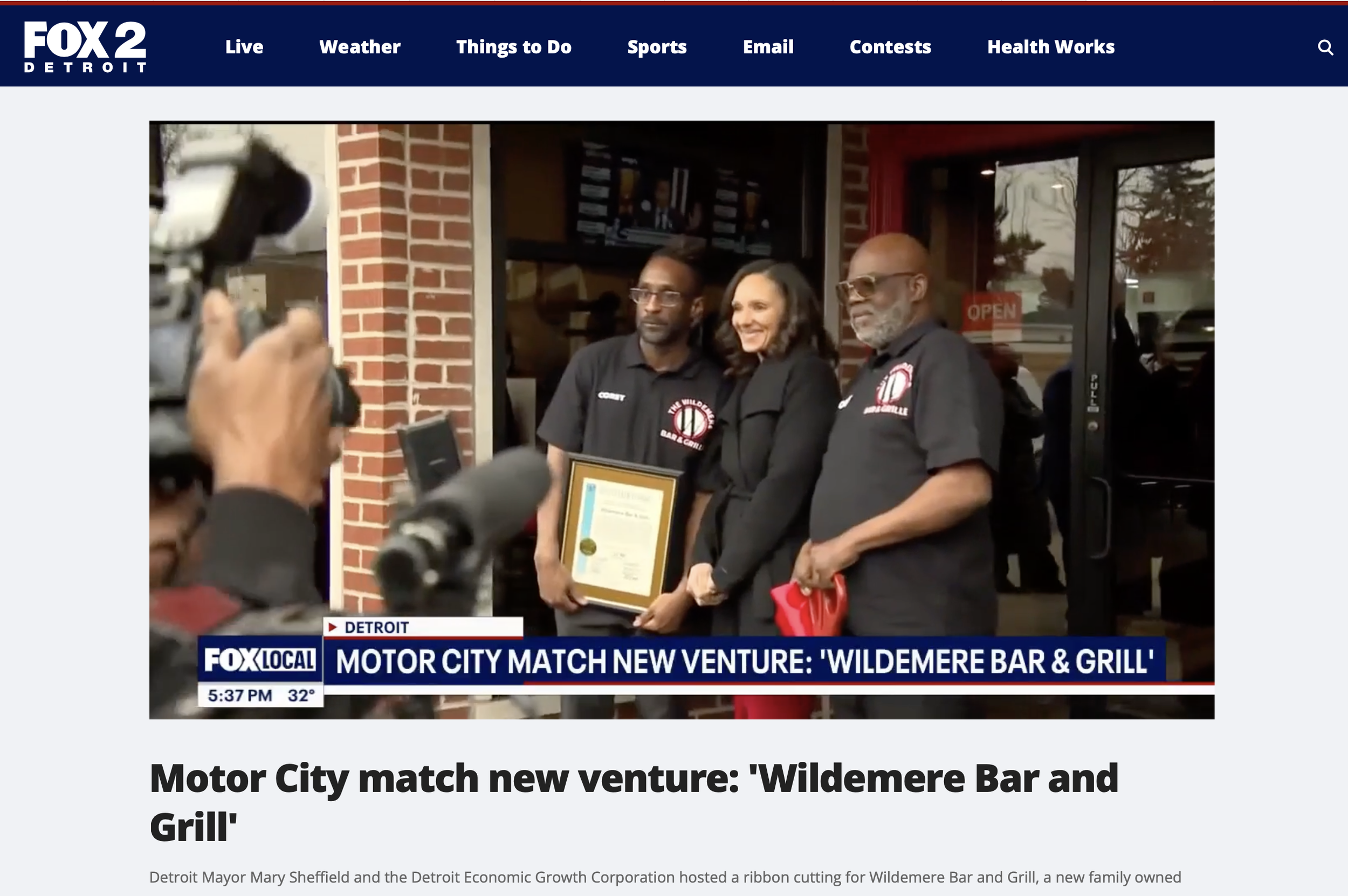 Group of three people standing outside a restaurant, two men and one woman, posing for a photograph during a ribbon-cutting event. One man is holding a framed certificate, and the woman is smiling. The restaurant has a brick exterior and a glass door with an 'OPEN' sign.