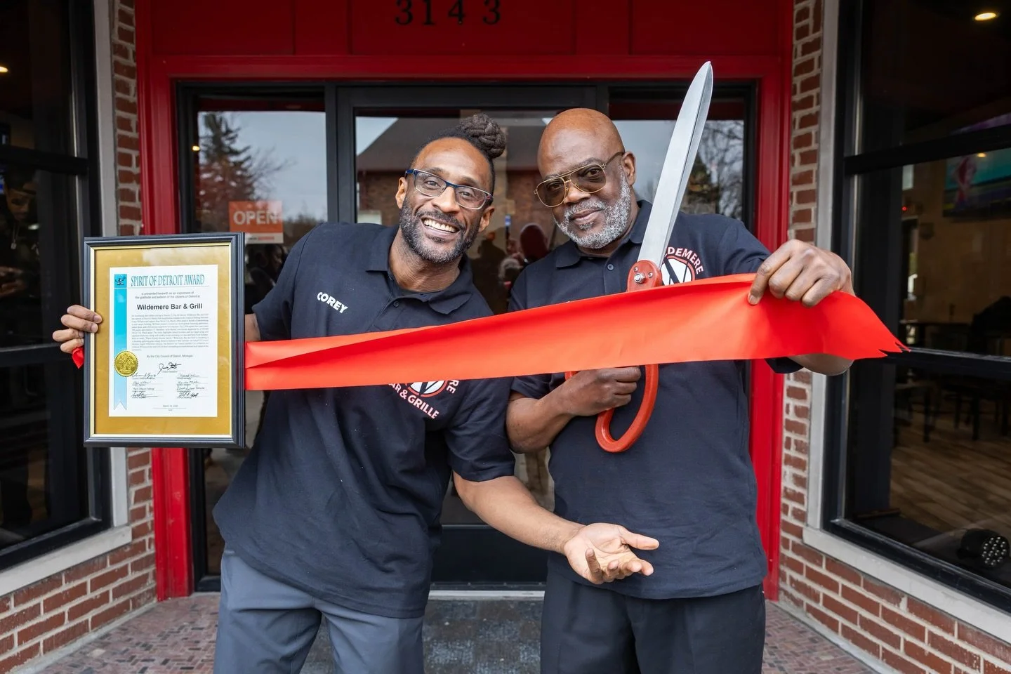 Two men stand outside a restaurant holding a framed award and a red ribbon with scissors. One man holds the award with a smile, while the other holds a pair of scissors ready to cut the ribbon. Both men are wearing black shirts with logos, and the ba