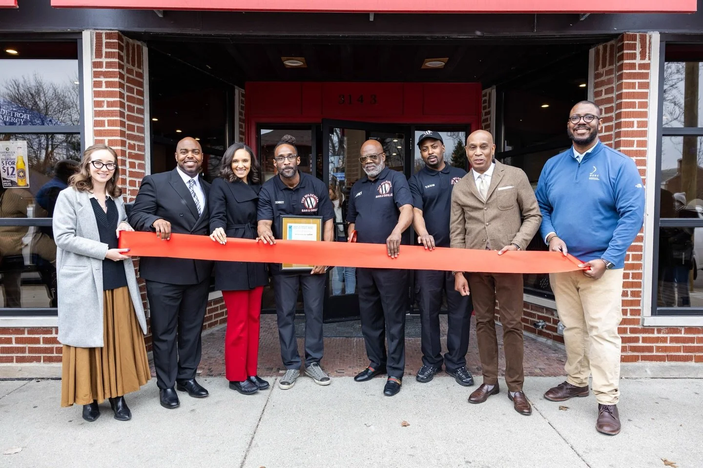 Group of nine diverse people, dressed in business and casual attire, holding a red ribbon in front of a building, celebrating a ribbon-cutting event.
