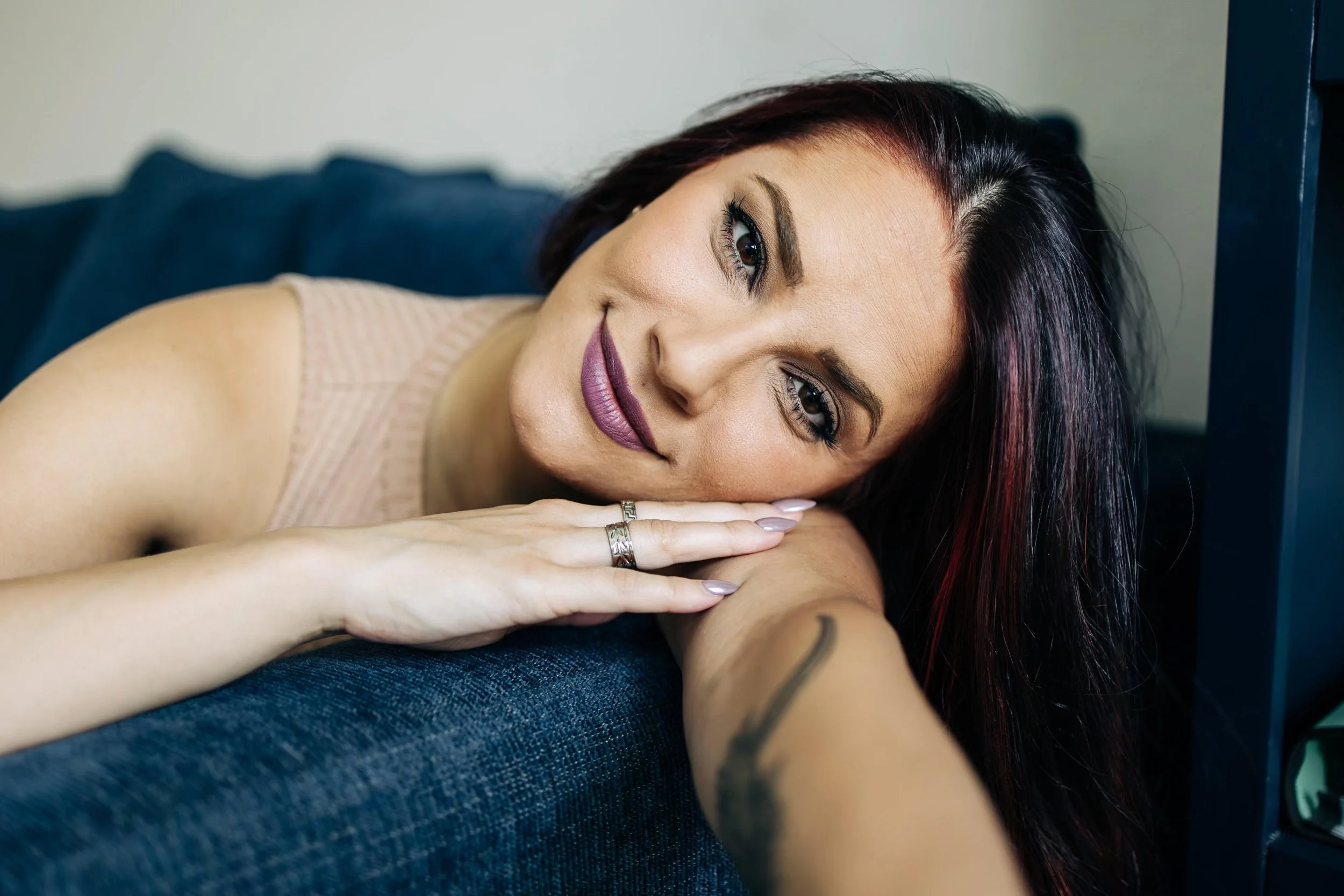 A woman with dark hair and makeup, resting her head on her forearms and smiling at the camera.