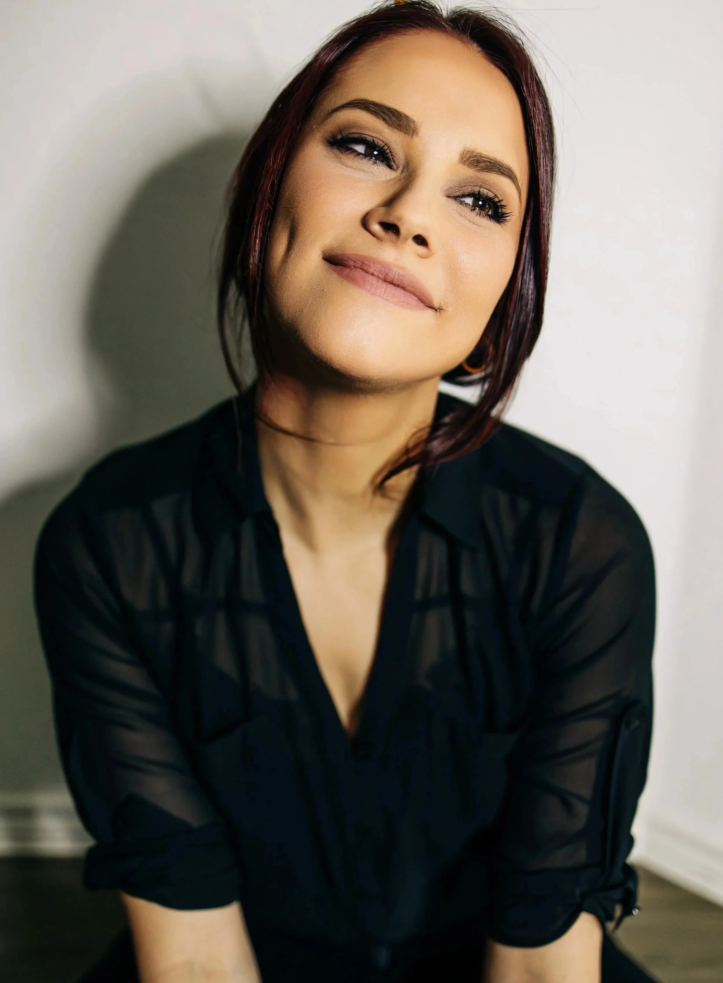 A woman with dark red hair wearing a black shirt, smiling softly, against a plain light background.
