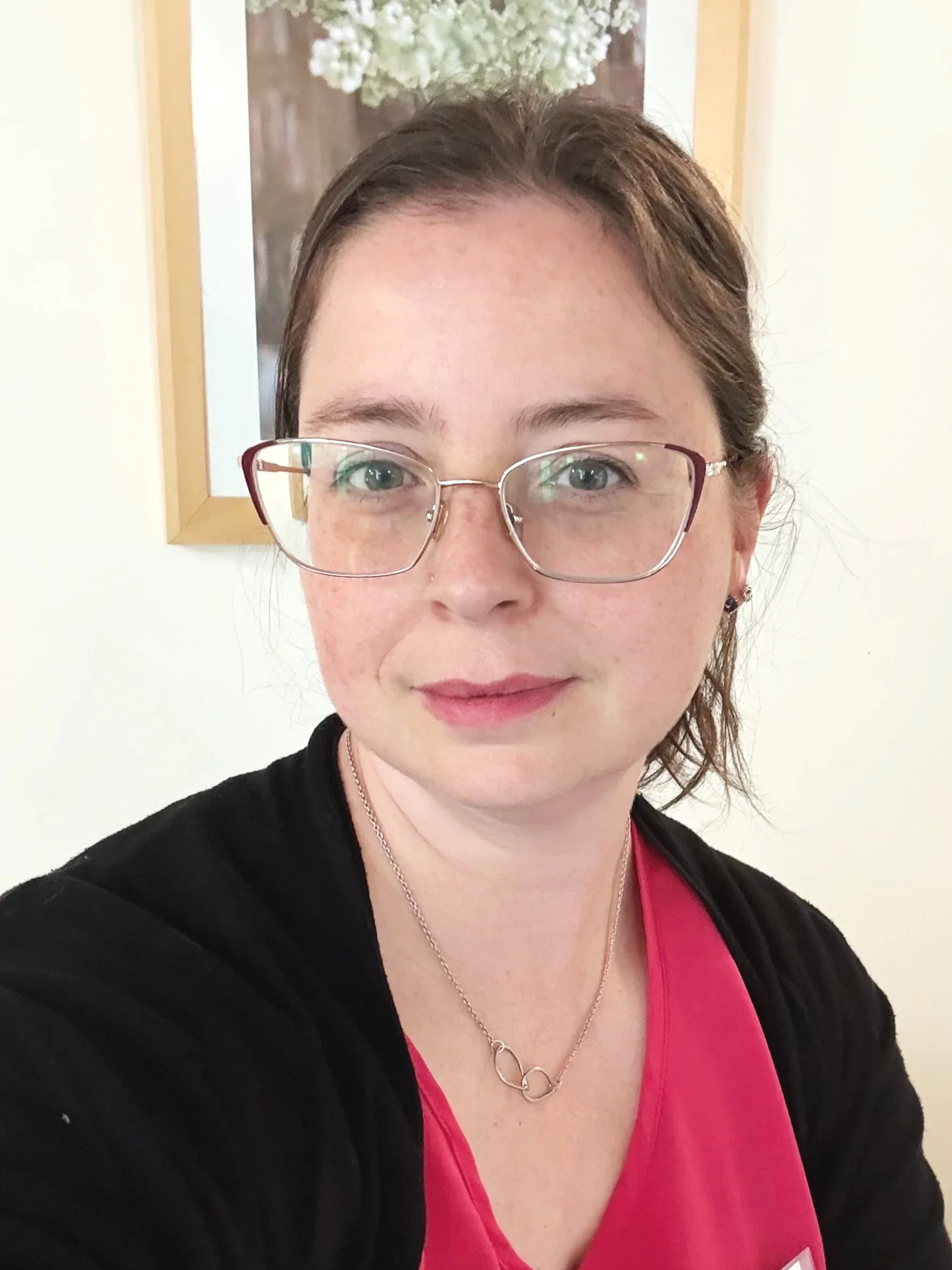 A woman with glasses smiling, wearing a black jacket over a pink top, with a silver infinity necklace, standing in front of a wall with a framed picture of white flowers.