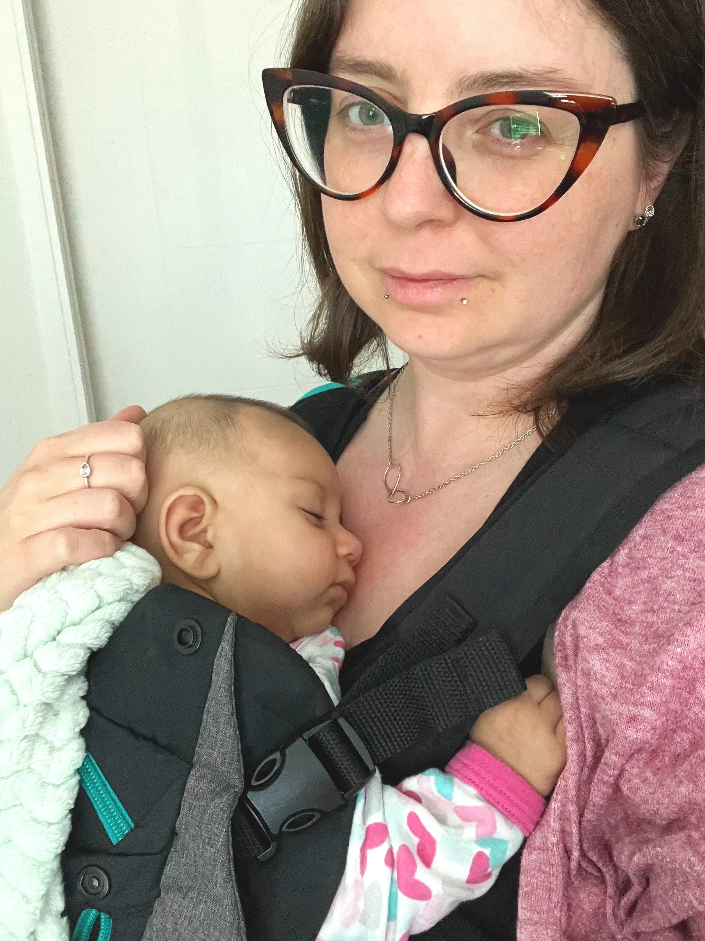 A woman with brown hair, wearing glasses, is holding a sleeping baby close to her chest. The woman has piercings on her lips and earrings, and is wearing jewelry, a black baby carrier, and a pink sweater. The baby is wrapped in a white blanket with pink, blue, and yellow patterns.