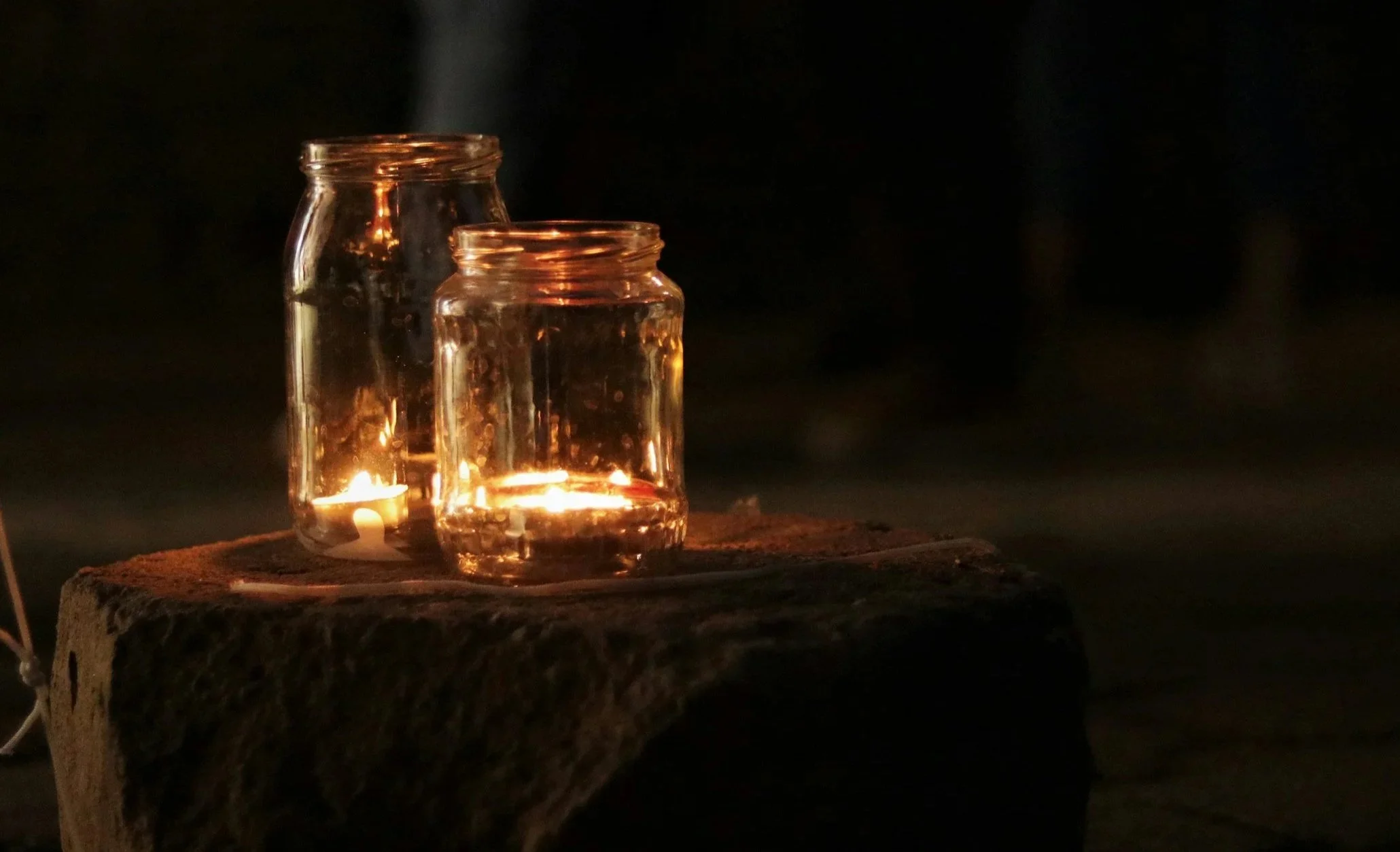 Two glass jars with candles inside placed on a brick surface against a dark background, with one jar emitting a candle flame and smoke.