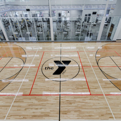 Showcasing a project by DOUBLEPLAY: Empty basketball court with the YMCA logo at center and elliptical gym equipment visible through glass wall behind.