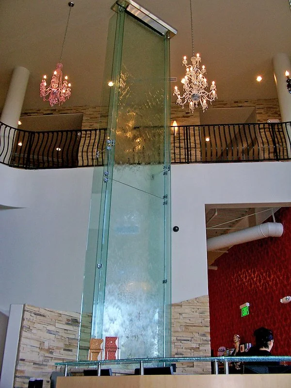 Showcasing a project by DOUBLEPLAY: Indoor space with a glass feature, a shaft extending through two levels, chandeliers hanging from the ceiling, a curved black railing on the upper level, and a decorative red wall