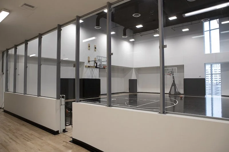 Showcasing a project by DOUBLEPLAY: An indoor basketball court enclosed by glass walls in a gymnasium with basketball hoops, black flooring, and high ceilings with skylights.