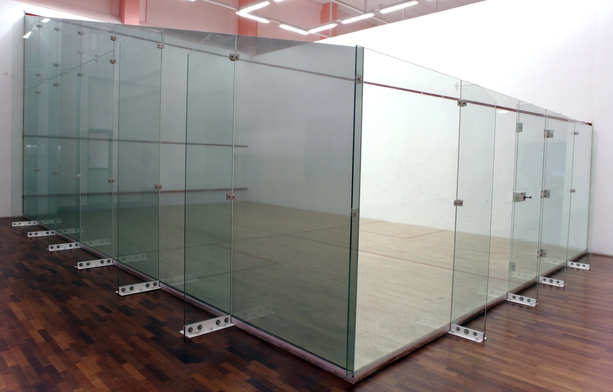 Showcasing a project by DOUBLEPLAY: A glass squash court with clear glass walls, wooden floor, and support brackets, located in an indoor sports facility.
