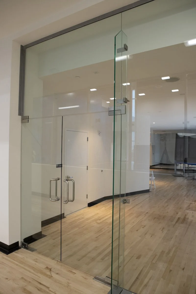 Showcasing a project by DOUBLEPLAY: Glass door with metal handles and hinges leading to a room with wooden flooring and white walls, illuminated by ceiling lights.