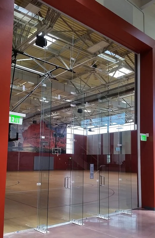 Showcasing a project by DOUBLEPLAY:View of an indoor basketball court through large glass doors. The court has wooden flooring, basketball hoops, and a red wall with a large red mural or decoration. Exit signs are visible on either side of the doors.