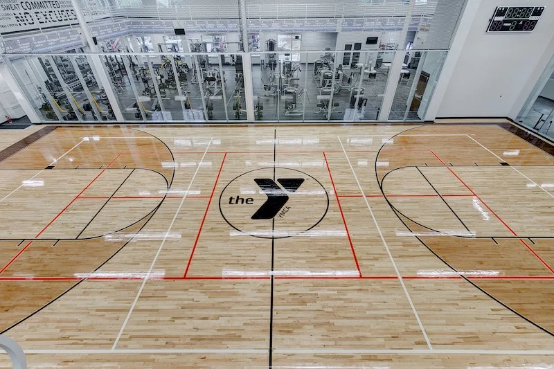 Showcasing a project by DOUBLEPLAY: Empty basketball court with a wood floor, the YMCA logo in the center, and an exercise room with equipment in the background.