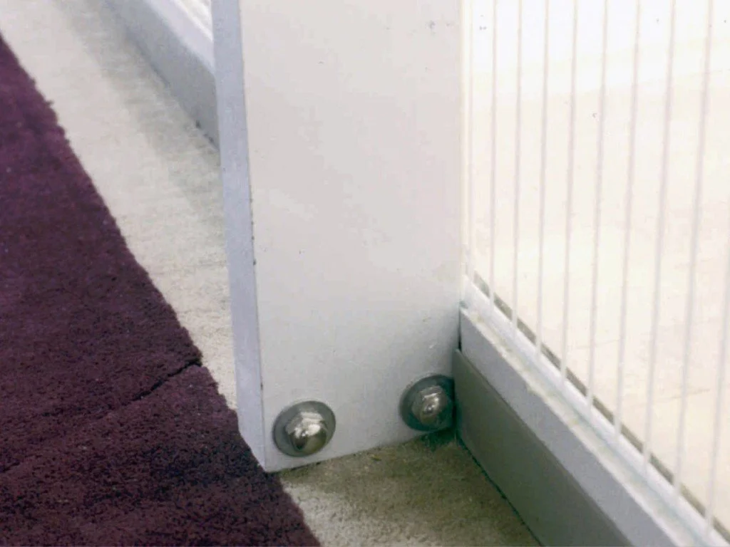 Design feature by DOUBLEPLAY: Close-up of the bottom corner of a piece of furniture or appliance with two visible metal fasteners, at the junction of a beige carpet, a purple rug, and a wall with vertical white paneling.