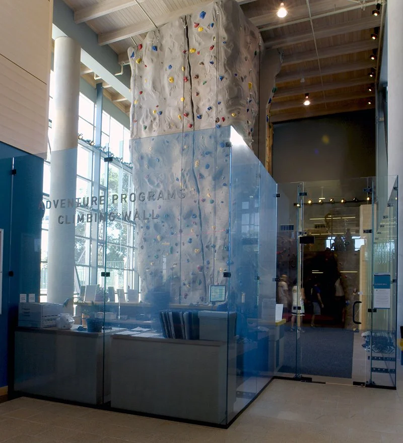 Showcasing a project by DOUBLEPLAY: Indoor climbing wall with colorful handholds and footholds, marked as Adventure Program Climbing Wall, enclosed in glass at an indoor facility.