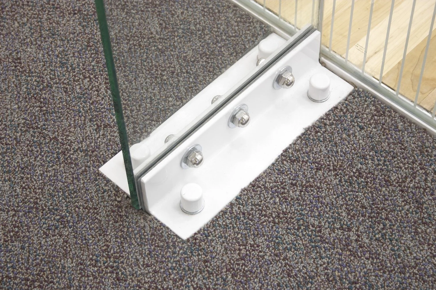 Design feature by DOUBLEPLAY: Close-up of a metal and glass bracket with a white base, four screws, two white knobs, and a glass panel, placed on speckled brown and gray carpet, with a background of boxed wooden panels and metal framing.