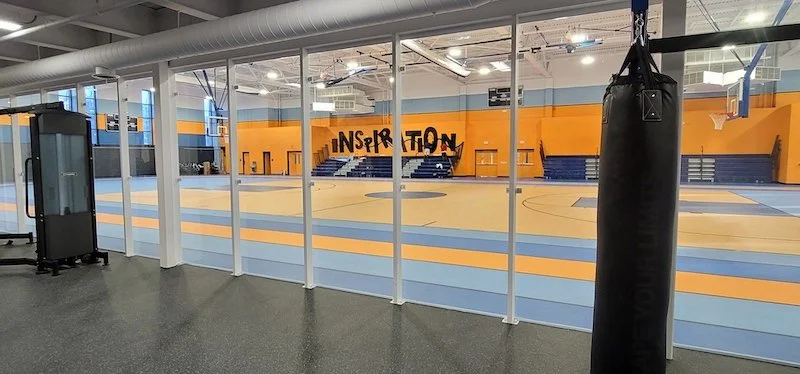 Showcasing a project by DOUBLEPLAY: View of an empty indoor basketball court seen through a glass window, with a punching bag hanging on the right and the word 'INSPIRATION' written on the wall at the far end.