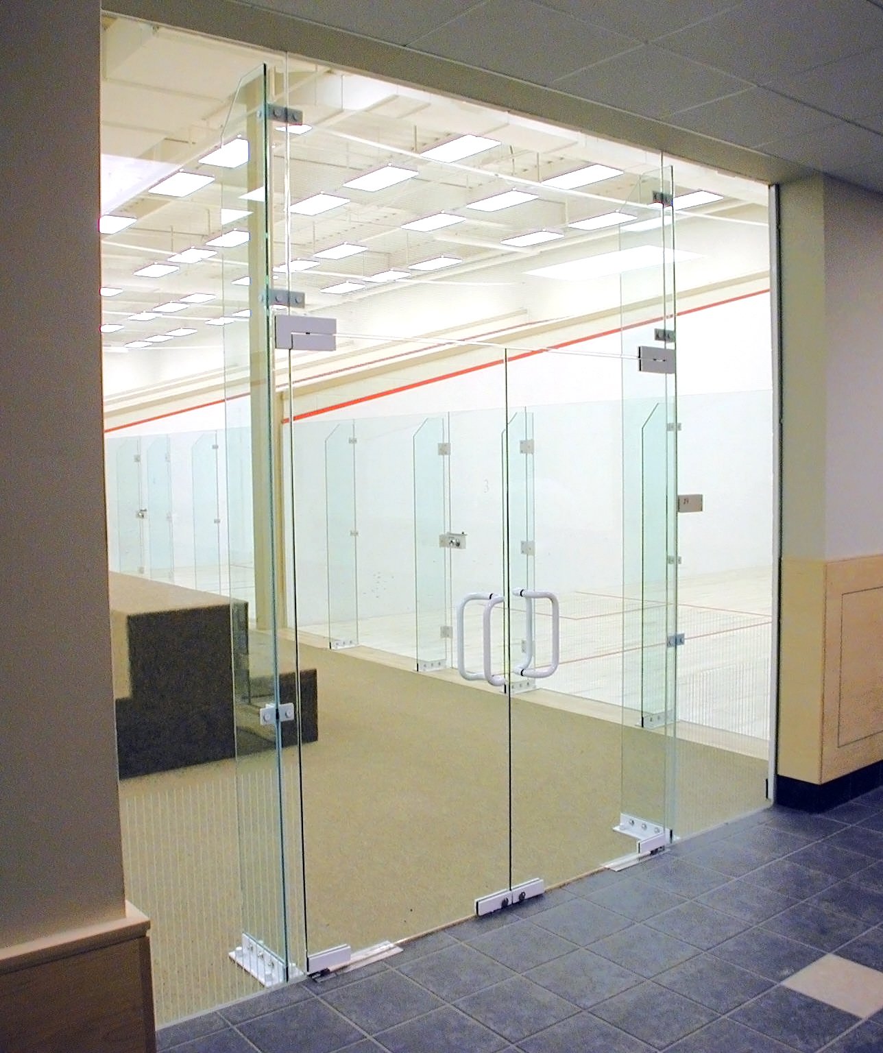 Showcasing a project by DOUBLEPLAY: Empty indoor sports court with glass walls and doors, overhead lighting, and seating area.