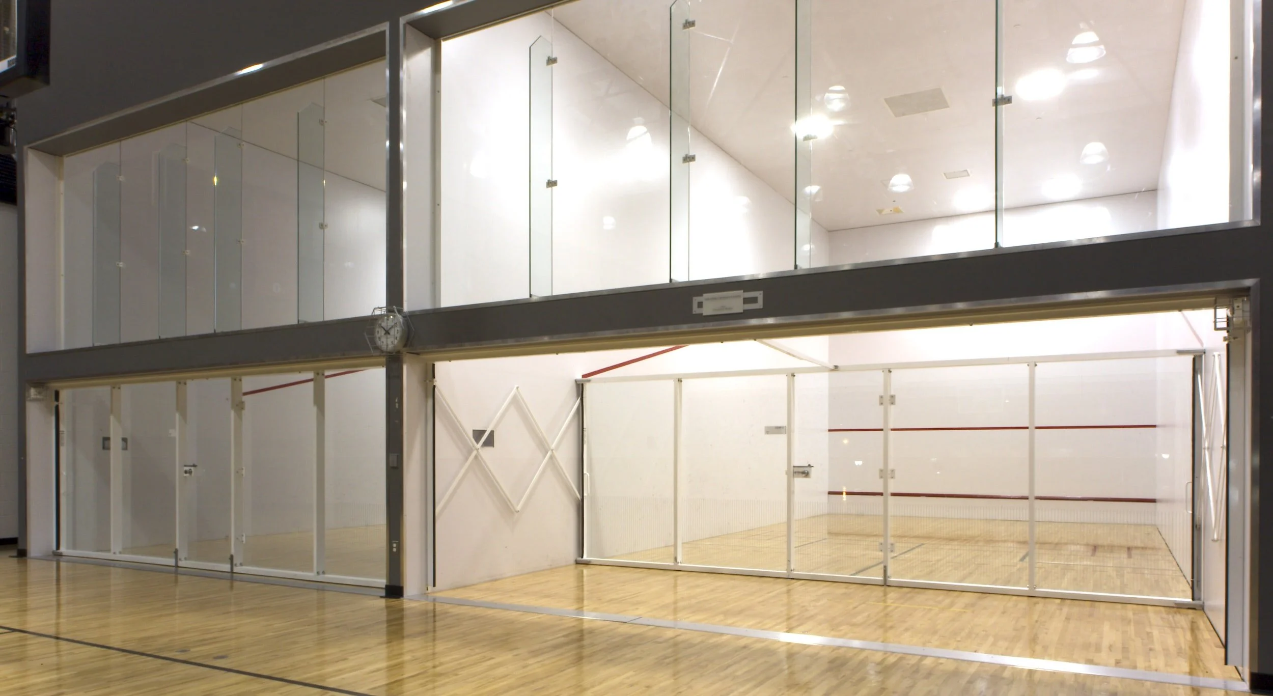 Showcasing a project by DOUBLEPLAY: Empty indoor squash court with glass walls, wooden floor, and bright overhead lighting.