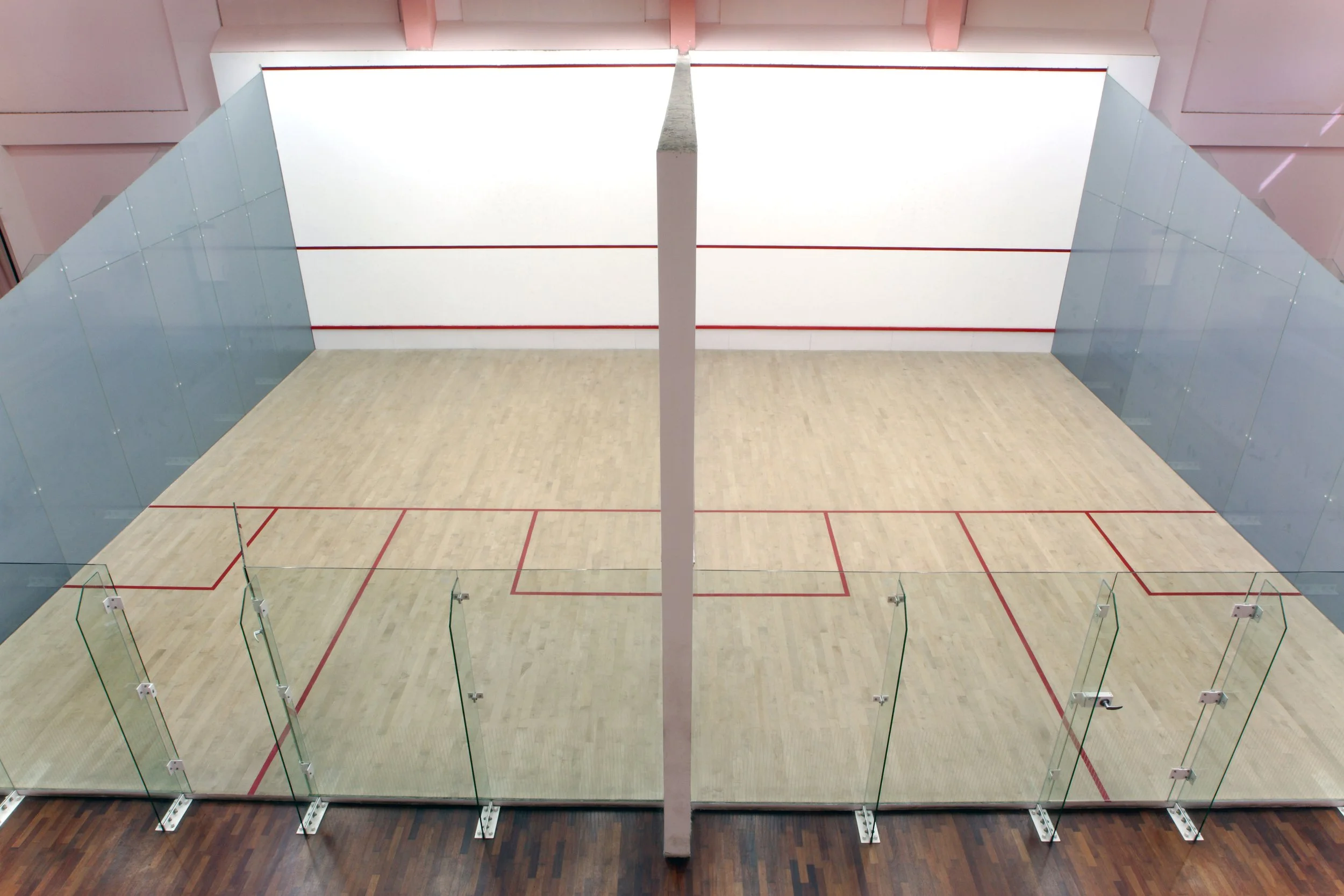 Showcasing a project by DOUBLEPLAY: An indoor squash court with glass walls, wooden flooring, and net lines marked on the floor.