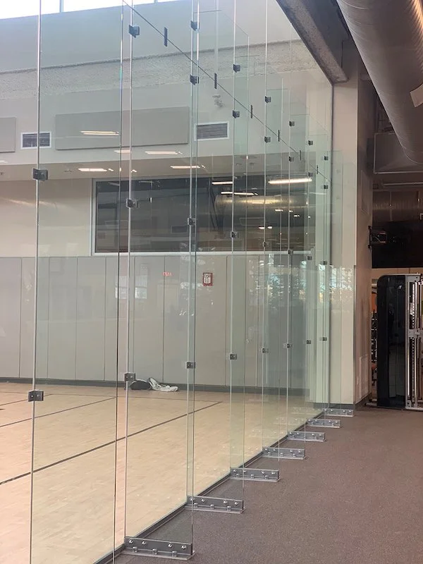 Showcasing a project by DOUBLEPLAY: Glass partition separating a gymnasium from a hallway, with a wooden sports floor on one side and gray carpet on the hallway side.