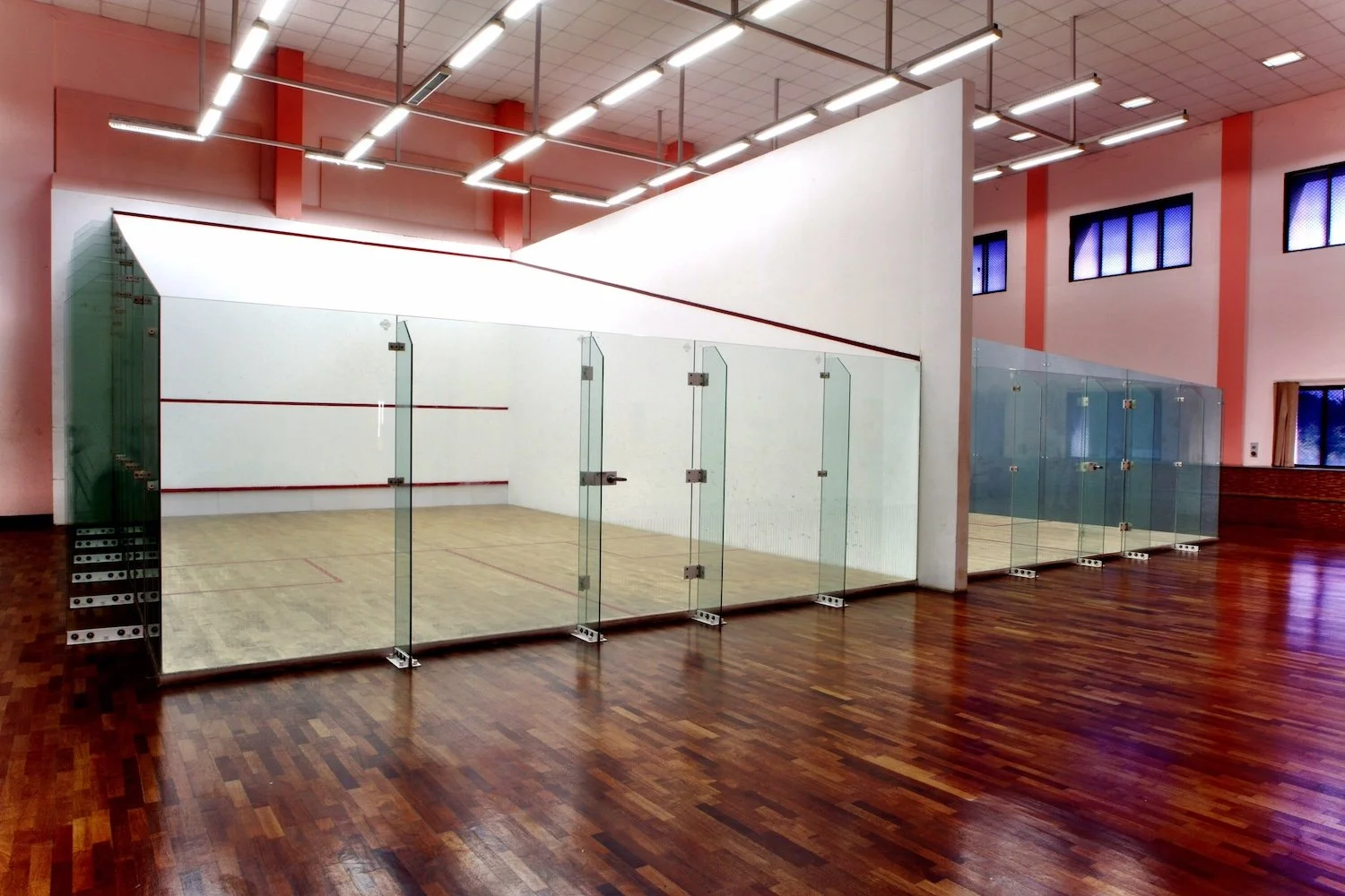 Showcasing a project by DOUBLEPLAY: An indoor squash court with glass walls and wooden flooring, lit by ceiling lights, located in a gymnasium with pink walls and windows.