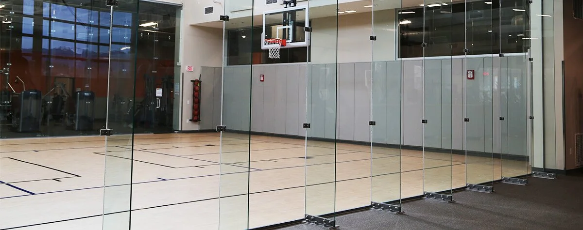 Showcasing a project by DOUBLEPLAY: An indoor basketball court visible through glass walls in a gym, with a basketball hoop and a fire extinguisher on the wall.
