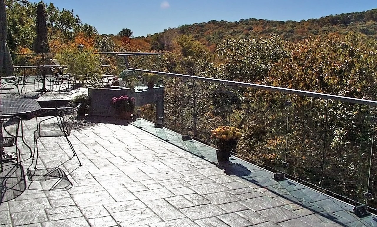 Showcasing a project by DOUBLEPLAY: A spacious outdoor terrace with stone pavement, surrounded by a glass railing and overlooking trees and a distant hillside. There are metal chairs and tables on the terrace, along with potted flowers.