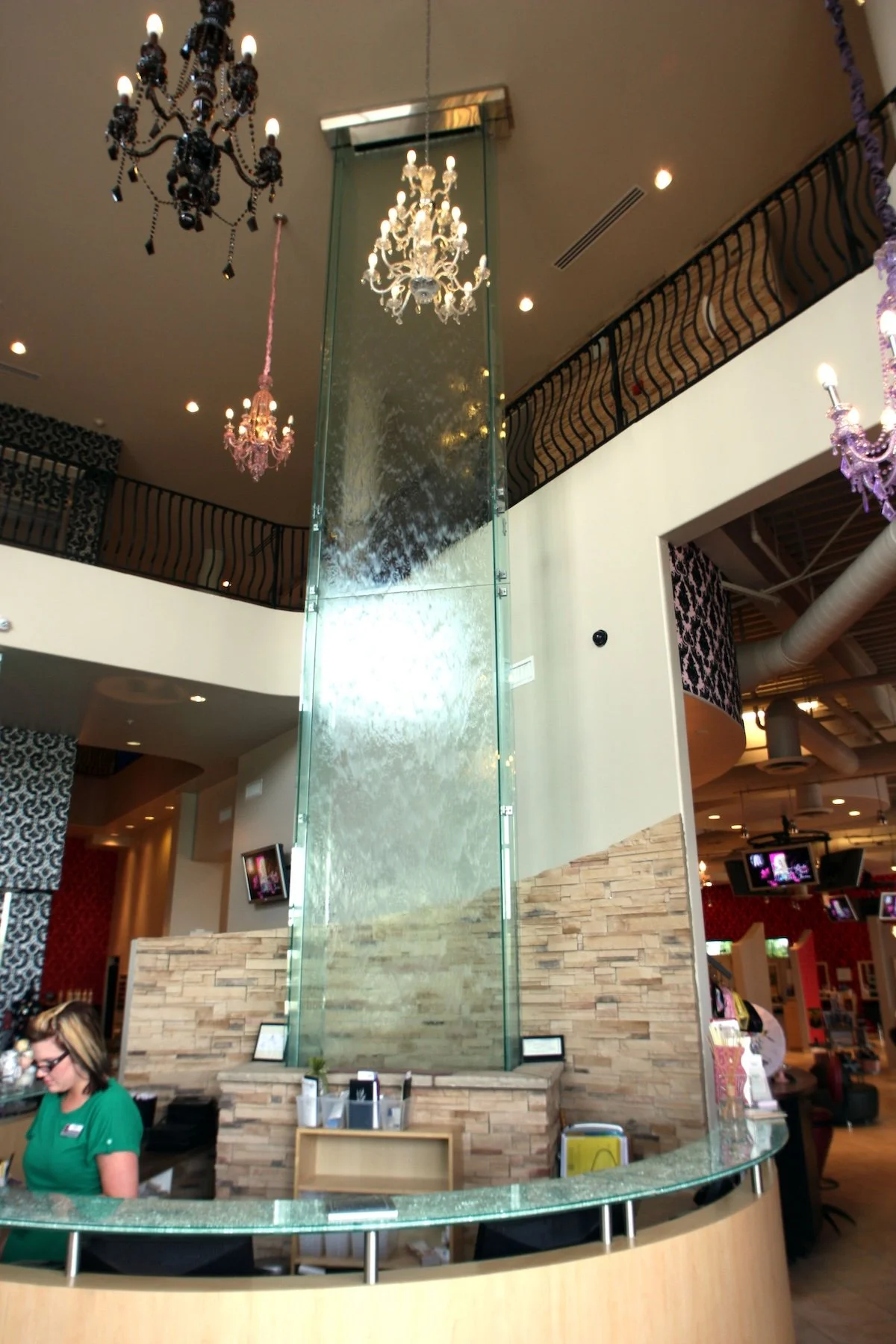 Inside a hotel or restaurant with chandeliers hanging from the ceiling, a glass water feature or decorative panel, a reception desk, and a woman working behind the counter.