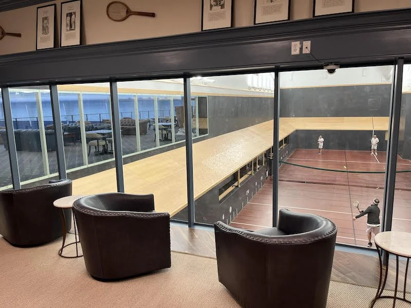 Showcasing a project by DOUBLEPLAY: Indoor tennis court viewed from a lounge with three black armchairs and small round tables.