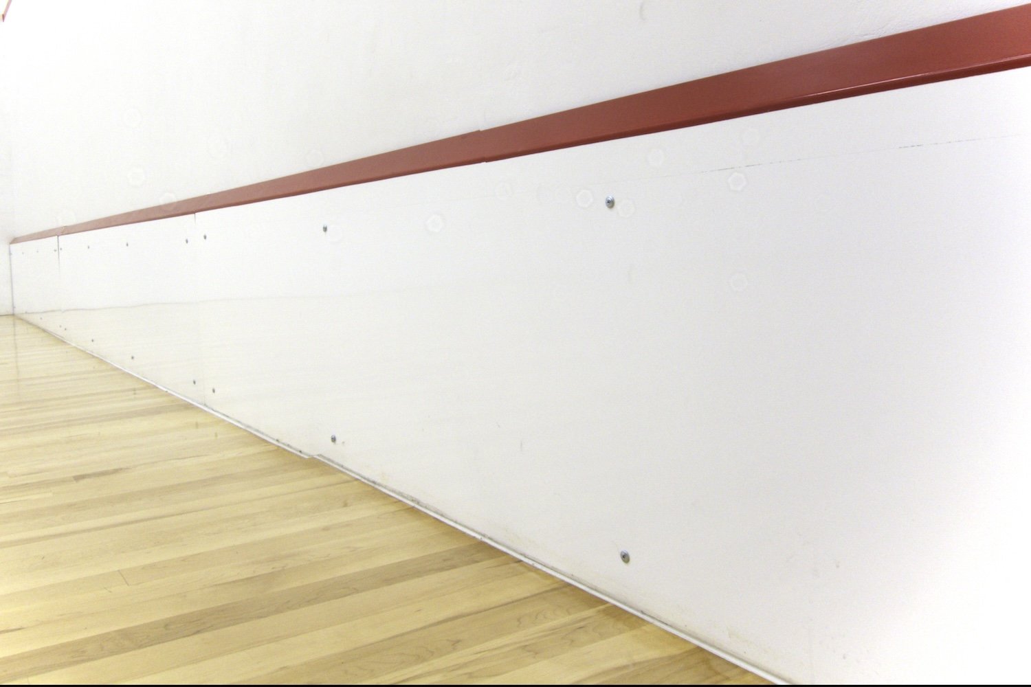 A wooden gym floor and a white wall with a brown handrail at the top, seen from a low angle.