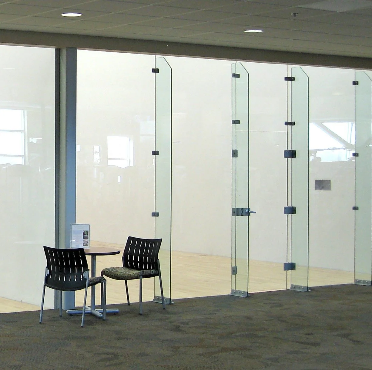 Showcasing a project by DOUBLEPLAY: Interior view of a modern office with glass walls, a seating area with two black chairs and a small round table, and wood flooring visible through the glass.