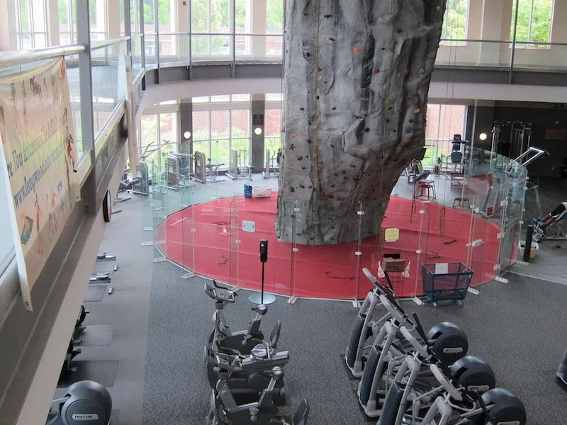 Showcasing a project by DOUBLEPIndoor gym featuring a large artificial rock climbing wall with a red padded floor for safety, surrounded by exercise equipment like ellipticals and stationary bikes, enclosed by glass walls and facing exterior windows.