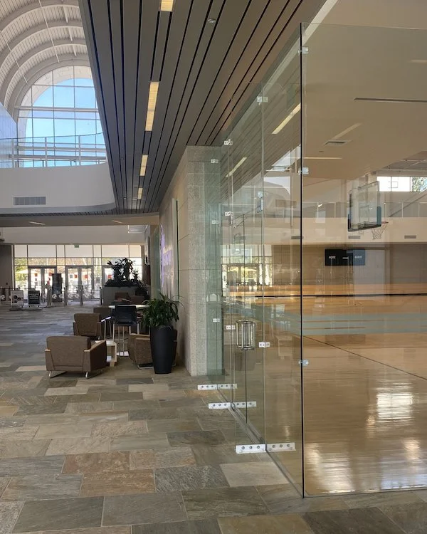 Showcasing a project by DOUBLEPLAY: Interior view of a modern, spacious gym or recreation center with a glass wall dividing a tennis or basketball court, seating area with chairs, plants, and a large window in the background.