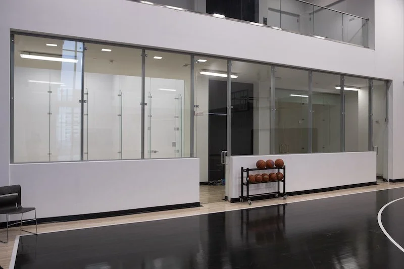 Showcasing a project by DOUBLEPLAY: Empty indoor basketball court with a small rack of basketballs, black and white walls, and glass-enclosed area.