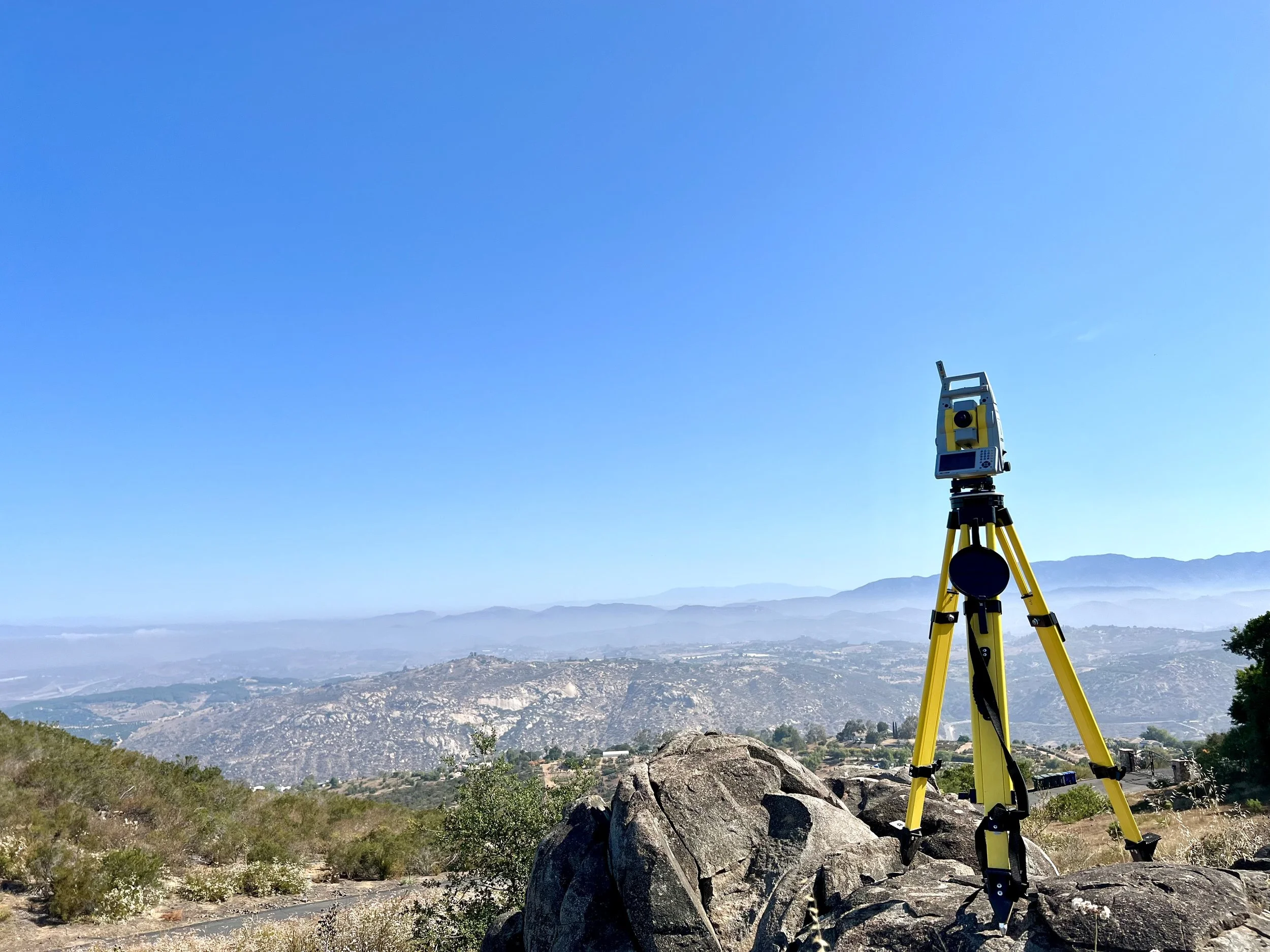 Surveying equipment on a tripod set up on rocky terrain overlooking a wide valley and mountain range under a clear blue sky.