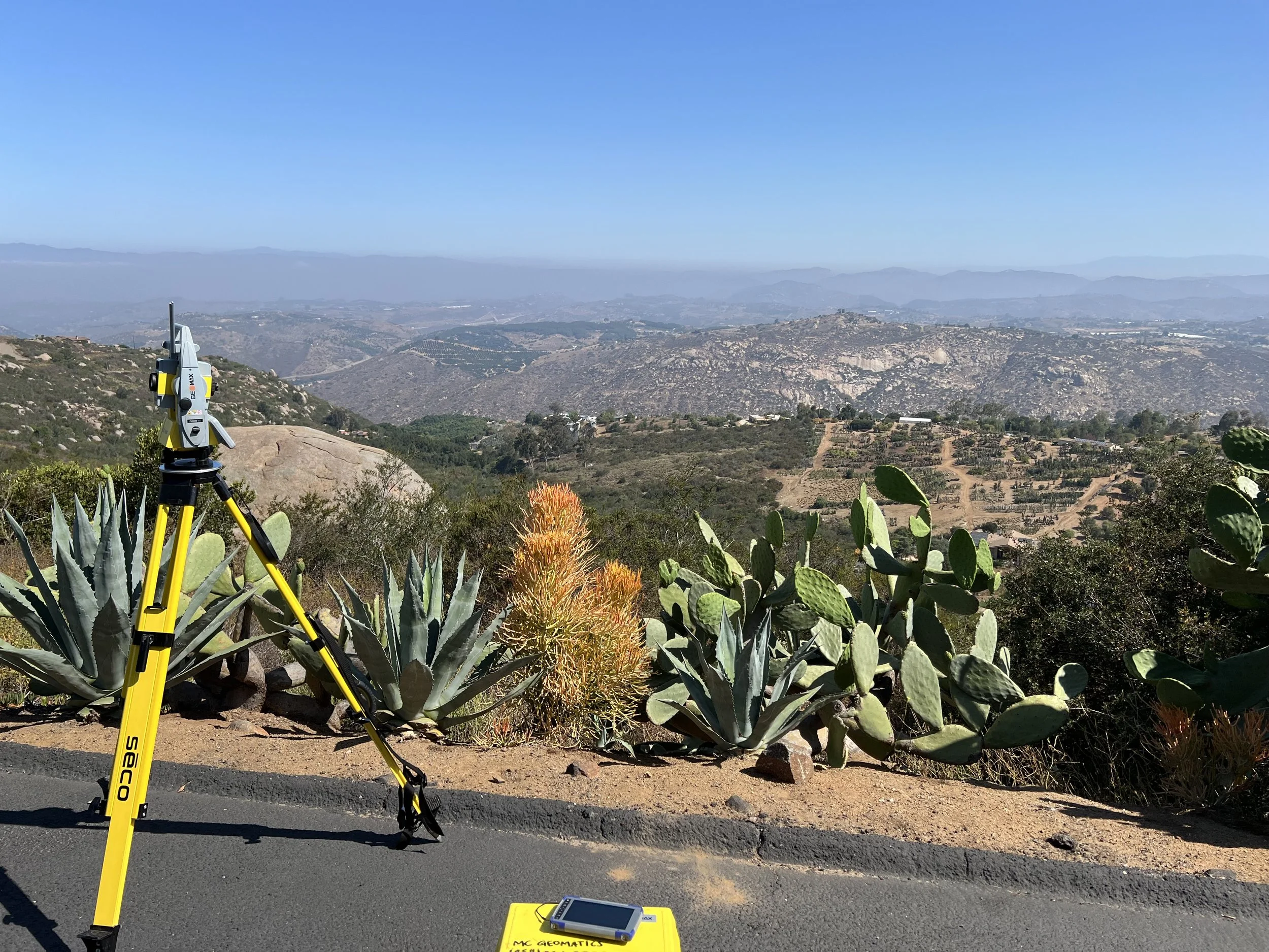 A surveying instrument set up on a tripod by the side of a paved road, with desert plants including cactus in the foreground, and rolling hills and mountains under a clear blue sky in the background.