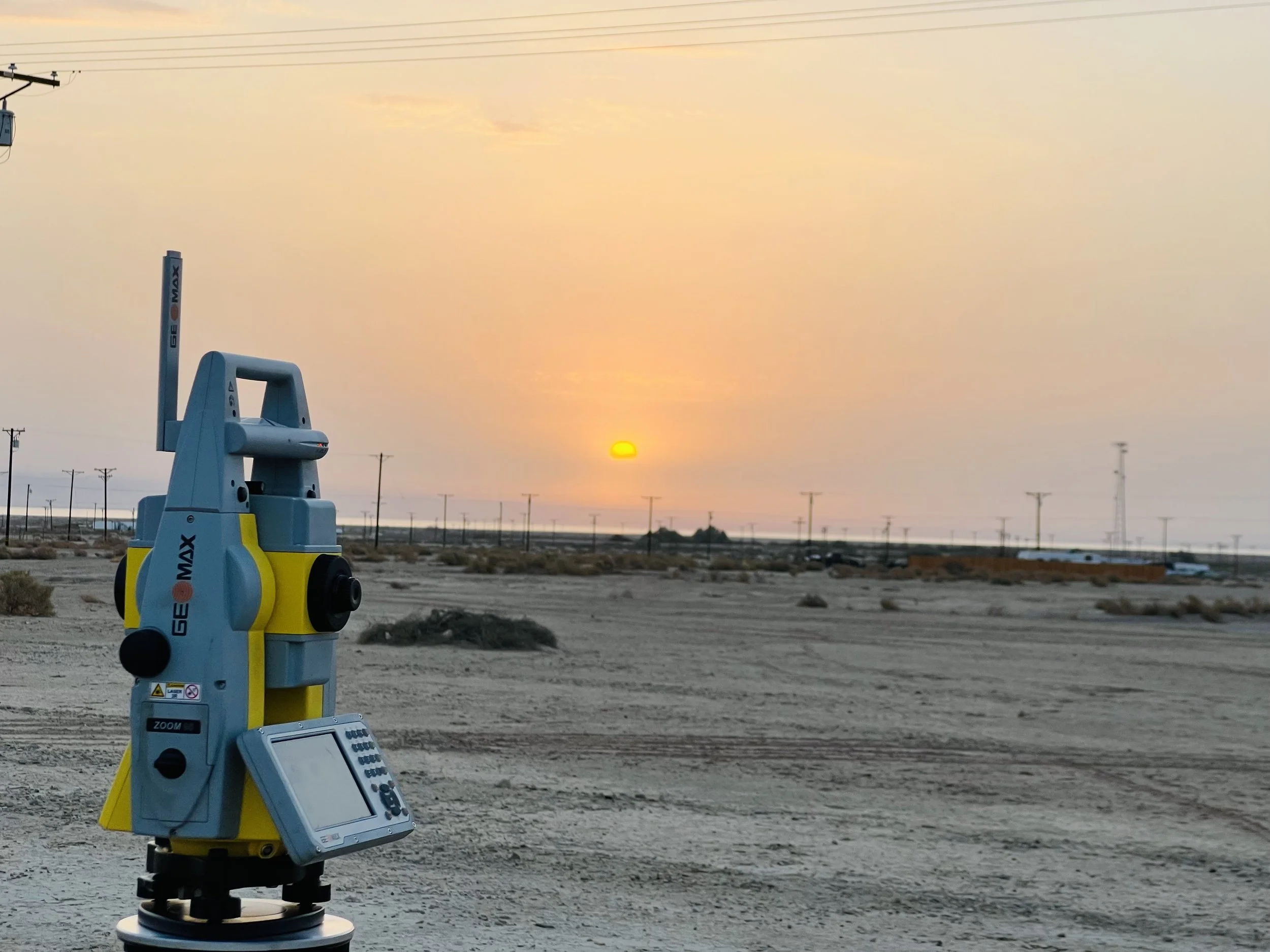 A surveying or mapping instrument mounted on a tripod in a desert landscape during sunset, with power lines and poles in the background.