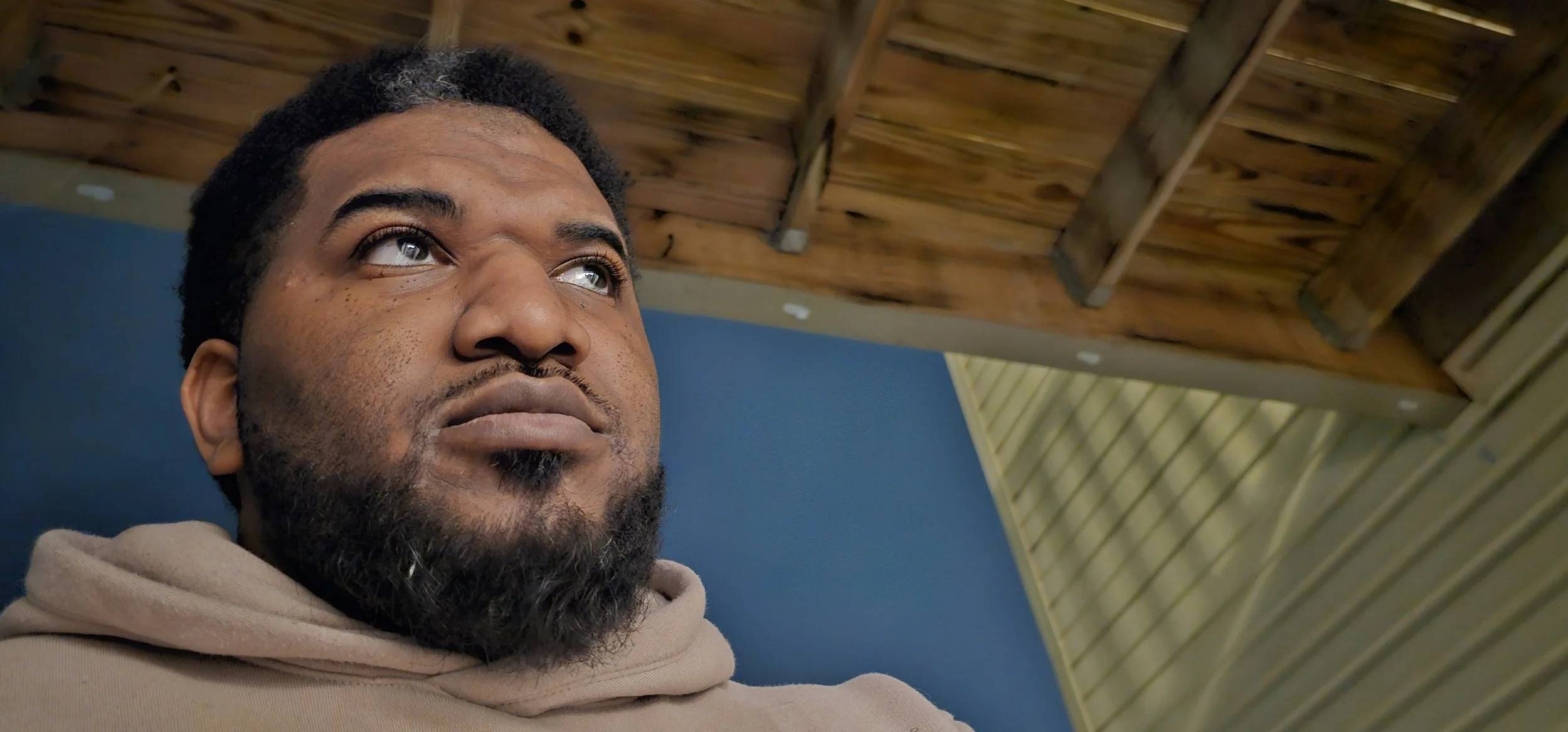 A close-up of a man with a beard and mustache, looking upward with a thoughtful expression, under a wooden roof with green and blue walls in the background.