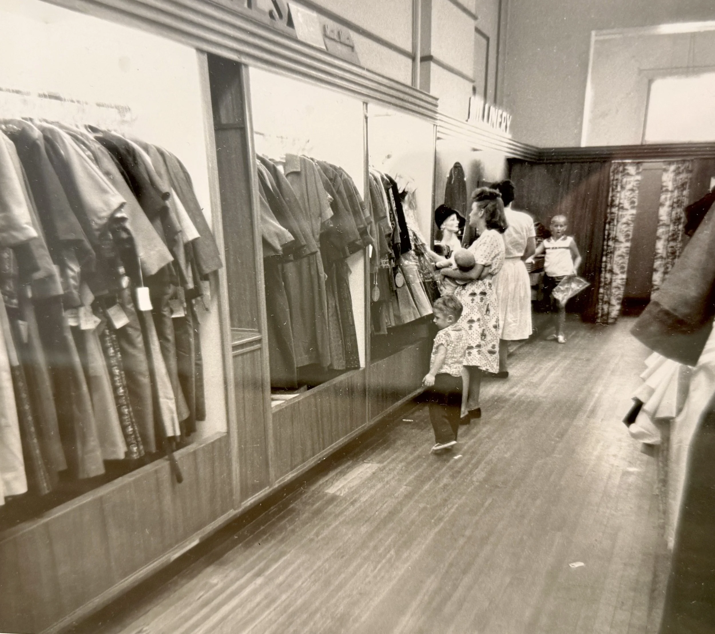 A vintage black-and-white photograph of a clothing store display featuring jackets and coats hanging on a wall-mounted rack, with people shopping and browsing in the background.