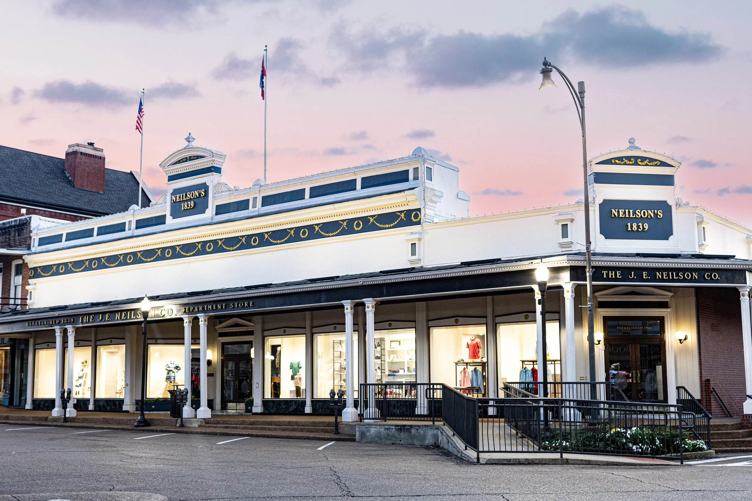 The corner of a historic department store called Neilson's, established in 1839, with a white facade, large windows displaying clothing, and American flags flying on top, under a pink and blue evening sky.