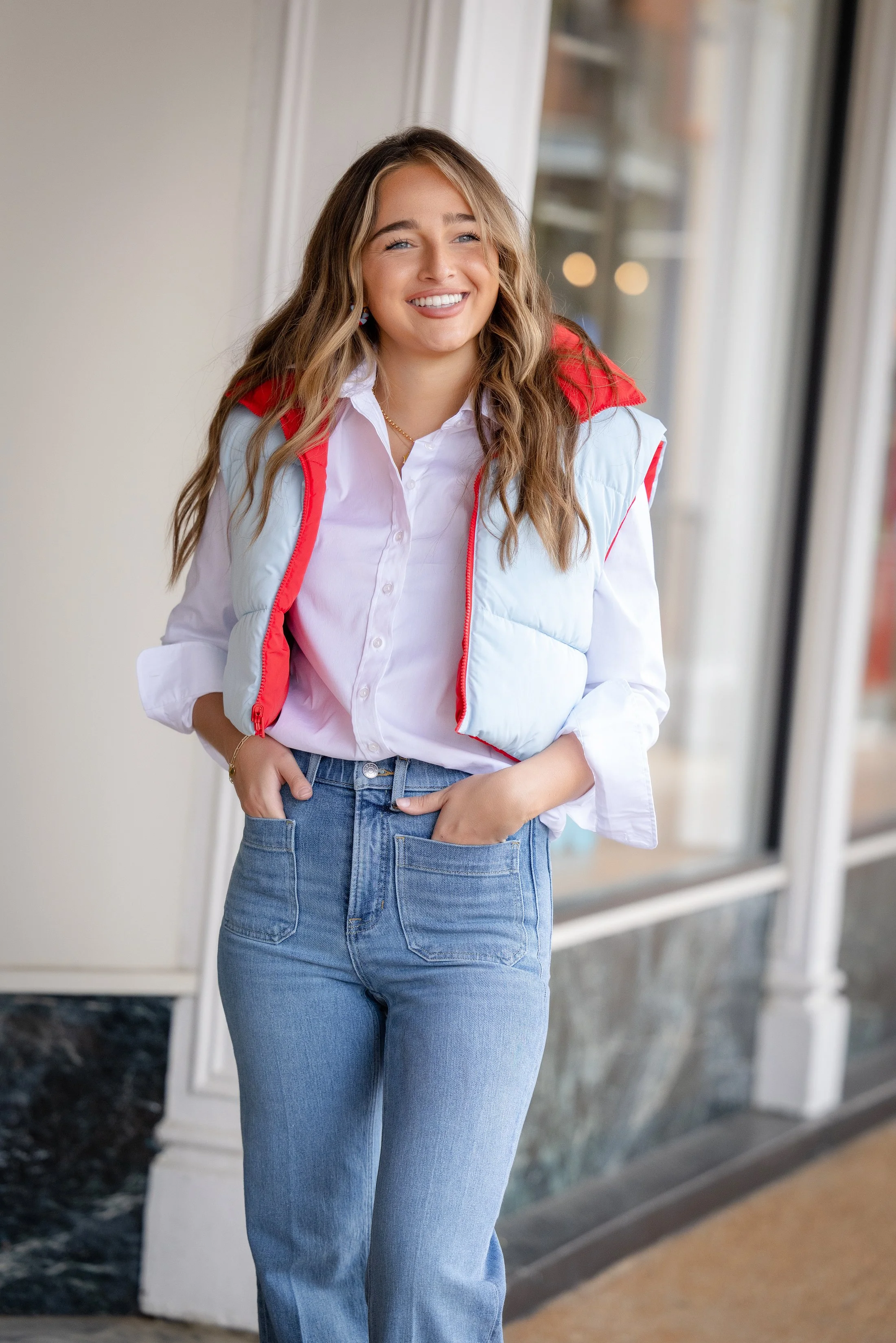 A young woman with long wavy blonde hair, wearing a white blouse, blue jeans, and a light vest with red accents, standing near a large window with a smile.