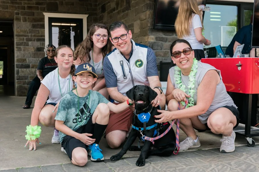 Group of six smiling people, including children and adults, gathered around a black service dog with a blue badge, outside a building with brick walls. Some are wearing leis, and one child is wearing a hat and a green tie-dye shirt.