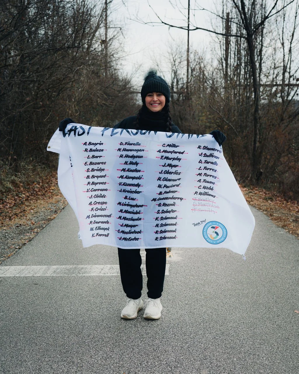 Person smiling outdoors holding a large white cloth with handwritten names, on a street with leafless trees.