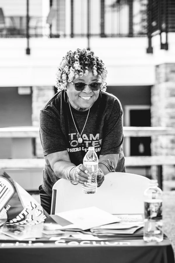 Smiling woman with curly hair, sunglasses, and jewelry holding a water bottle in a casual indoor setting.