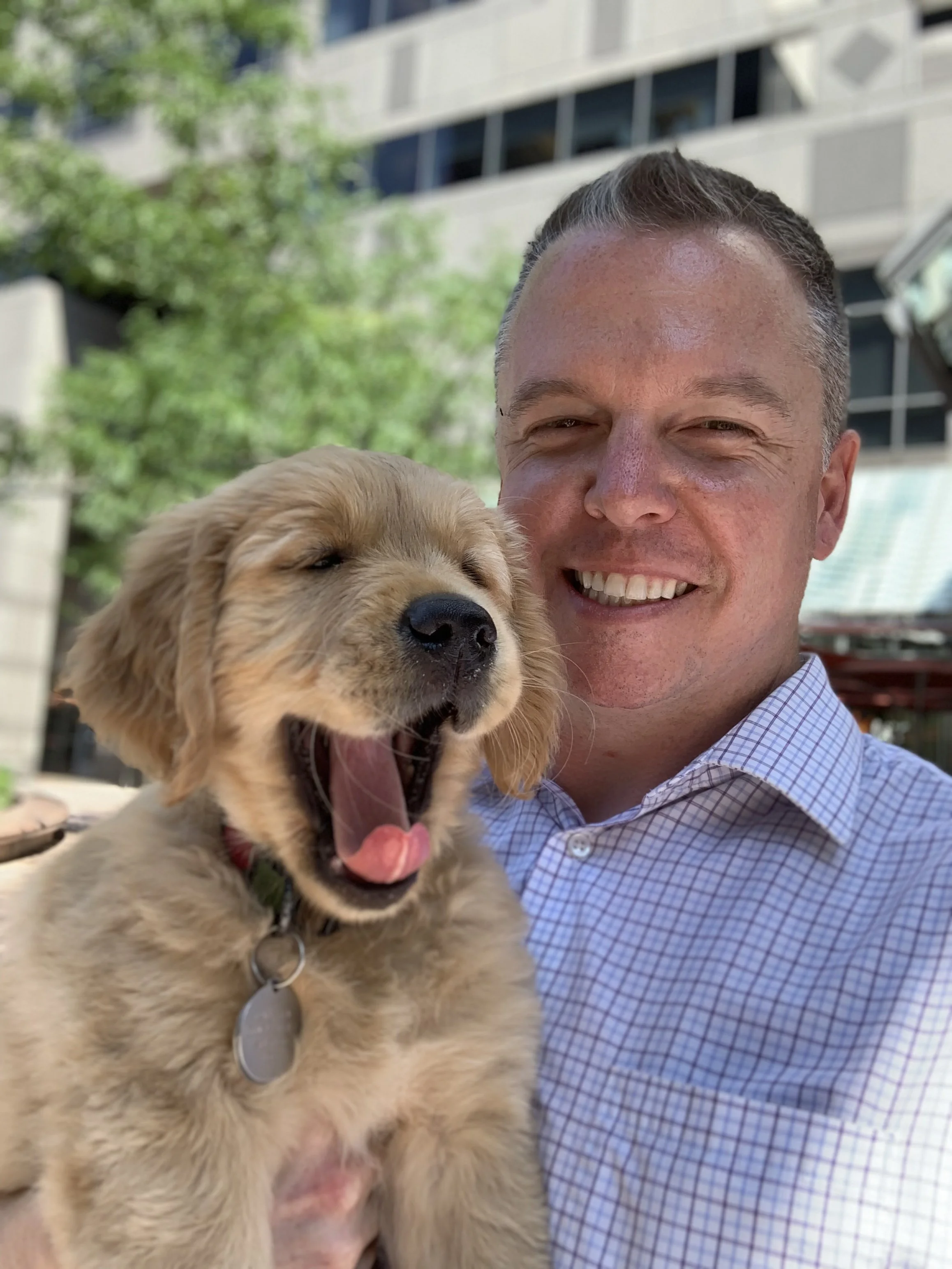 A man smiling at the camera holding a yawning golden retriever puppy outdoors.